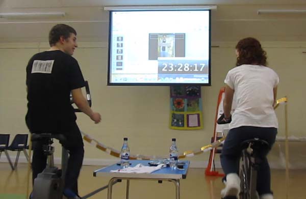A pair of cyclists viewed from the back, facing a giant screen showing a countdown timer